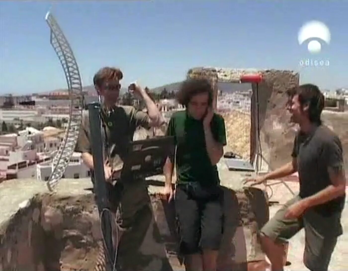 Three people on a rooftop beside a curved directional wifi antenna, town and sea behind.